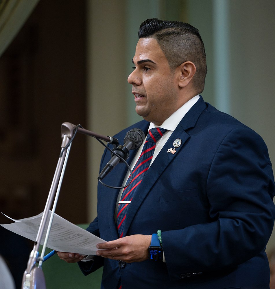 Asm. González standing at microphone, holding papers and speaking