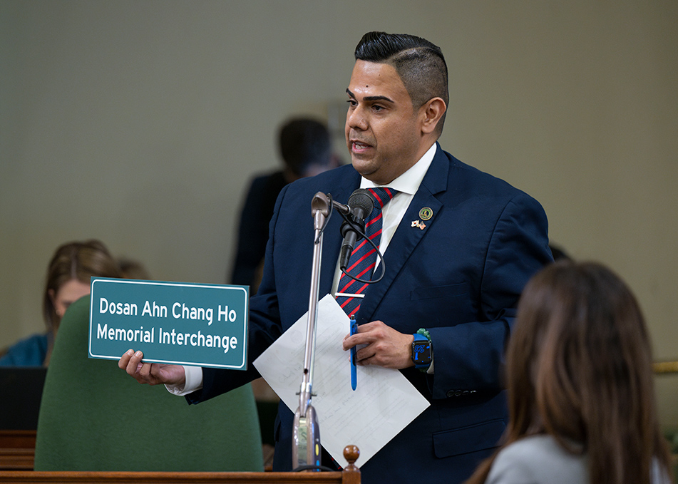 Asm. González standing at microphone, holding papers and a sign displaying Korean and English words