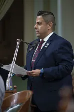 Asm. González standing at microphone, holding papers and speaking