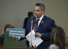 Asm. González standing at microphone, holding papers and a sign displaying Korean and English words