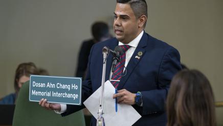Asm. González standing at microphone, holding papers and a sign displaying Korean and English words