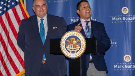 Former assemblymember Miguel Santiago at podium, speaking as Speaker Emeritus Pérez listens on