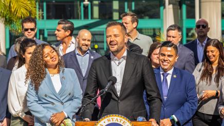 Asm. González listening on as Asm. Haney speaks at podium