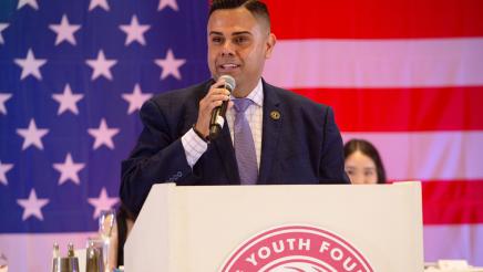Asm. González at podium, speaking, with large U.S. flag in background