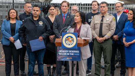 Guest speaker at podium, speaking, as Asm. González and others listen on