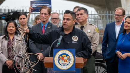 Asm. González at podium, speaking and holding tangled wires, as others listen on